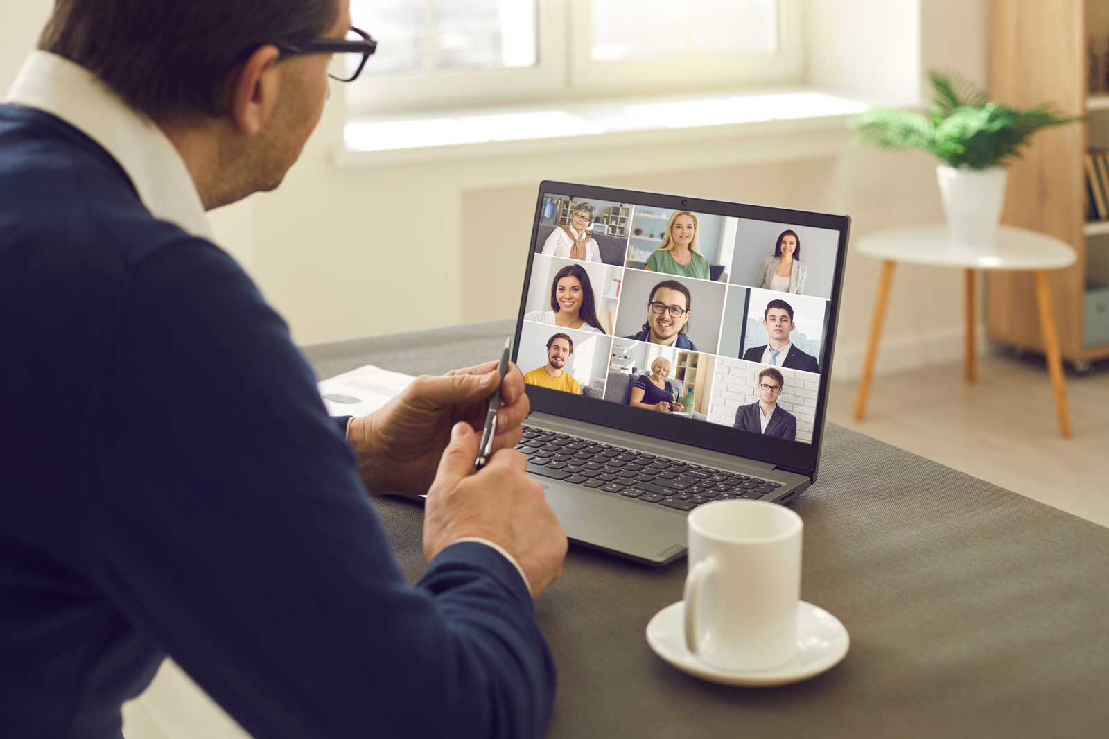 Eine Person sitzt am Tisch und betrachtet ein Videoanruf-Meeting auf ihrem Laptop mit mehreren Teilnehmern.